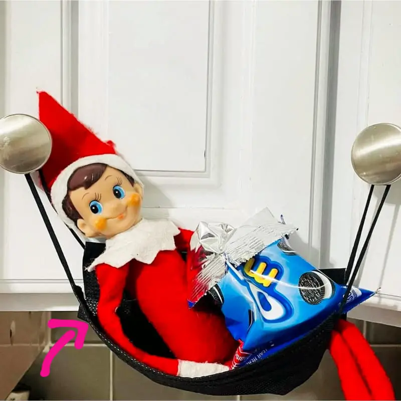Lazy Elf on the Shelf pose idea - last minute set up using a face mask hung on 2 kitchen cabinet knobs as a hammock for your Elf - with elf laying in it with a small back of cookies